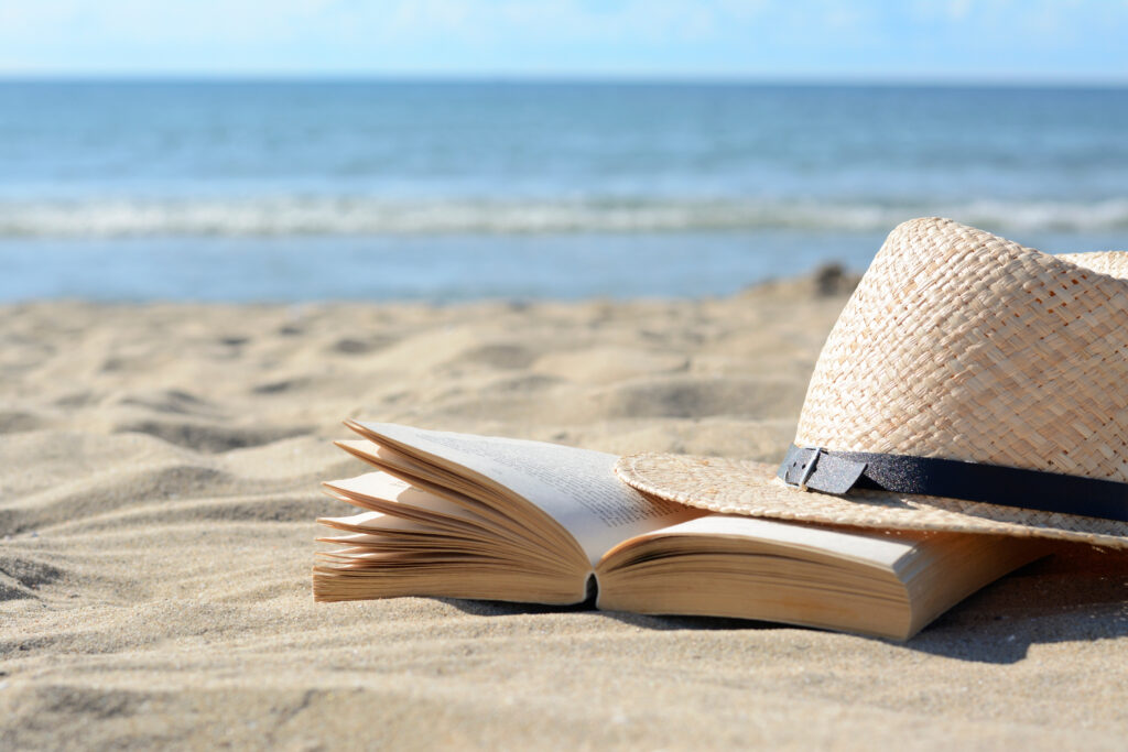 Offenes Buch und Strohhut im Sand am Strand mit Blick aufs Meer.
