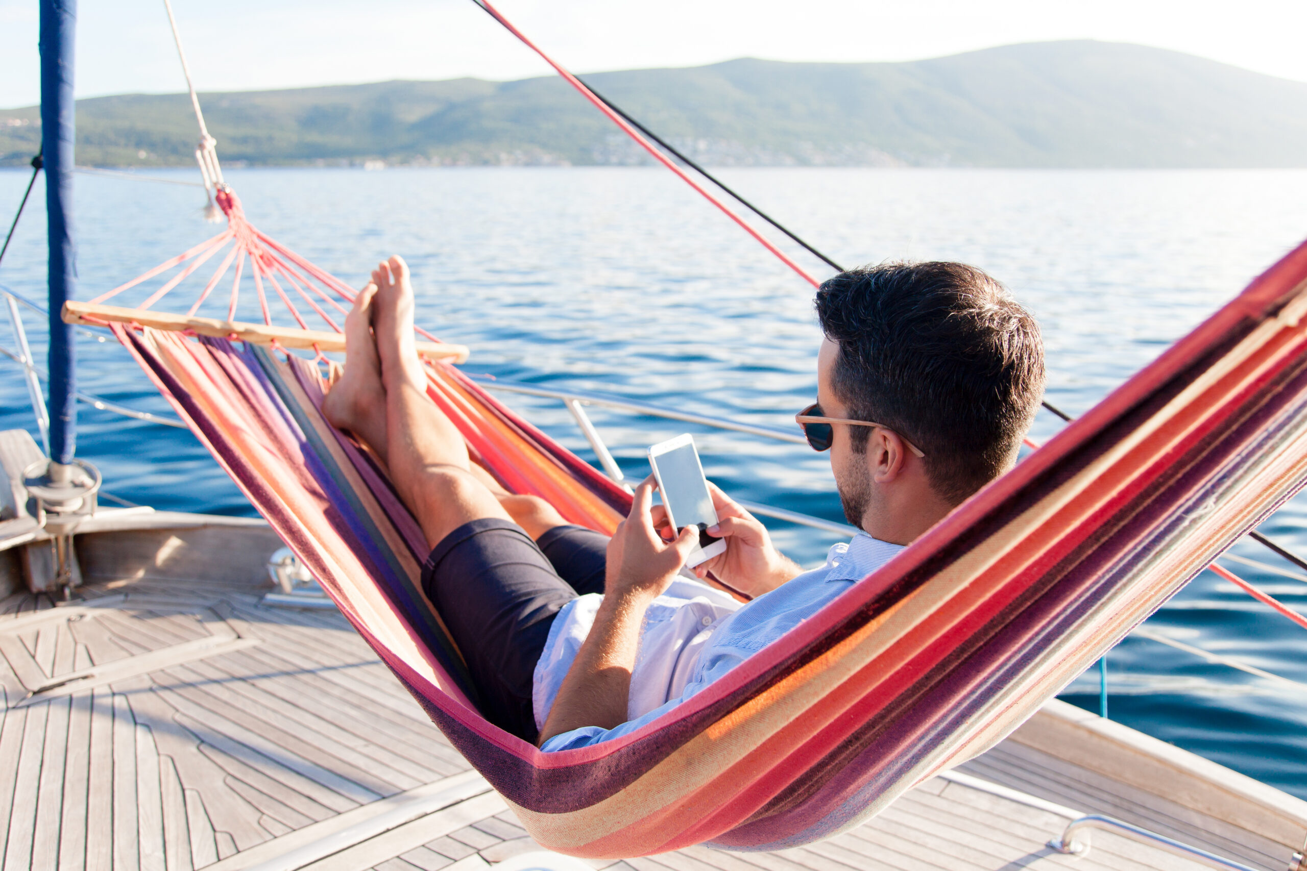 Man with mobile phone relaxing on yacht in hammock. Sea travel on sailboat. Traveler gaming on smartphone in summer vacation. Concept of doomscrolling, digital wellbeing, surfing the Internet, FOMO.