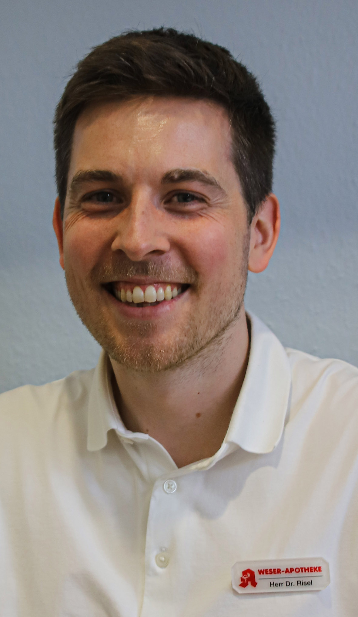 Apotheker Dr. Philipp Risel mit weißem Kittel, lächelt in die Kamera, im Hintergrund Regale mit Arzneimitteln.
